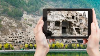 travel concept - tourist photographs caves on West Hill of Chinese Buddhist monument Longmen Grottoes (Longmen Caves, Dragon's Gate) from the east bank of the Yi River in spring season on tablet