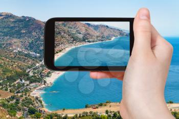 travel concept - tourist photographs Letojanni resort town of coast of Ionian Sea from Taormina city in Sicily Italy in summer on smartphone