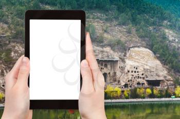 travel concept - tourist photographs Grottoes on West Hill of Chinese Buddhist monument Longmen Caves (Dragon's Gate) on bank of Yi River in China on tablet with cut out screen for advertising logo