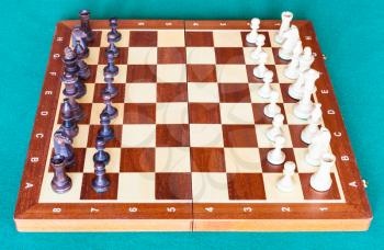 side view of wooden chessboard with chess pieces in starting position on green baize table