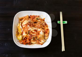 korean cuisine - top view of kimchi appetizer (spicy nappa cabbage) in white bowl and chopsticks on dark brown wooden table