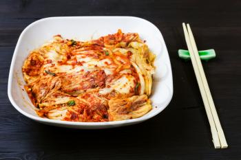 korean cuisine - kimchi appetizer (spicy nappa cabbage) in white bowl and chopsticks on dark brown wooden table
