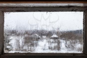 blurry view of russian village through water drops from melting ice on surface of frozen window of rural house in cold winter day (focus on surface of the glass)