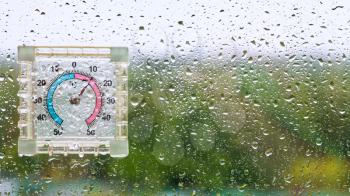 raindrops and outdoor wet thermometer on glass of home window and blurred city park on background on cool rainy autumn day