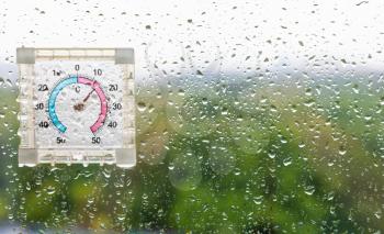 rain drops and outdoor thermometer on window glass and blurred woods on background on cool rainy autumn day