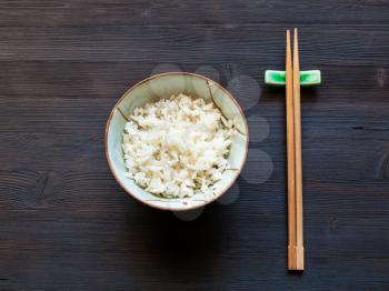 top view of portion of boiled rice in bowl and chopsticks on rest on dark wooden desk