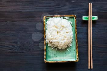 top view of portion of boiled rice on green plate and chopsticks on rest on dark wooden desk with copyspace