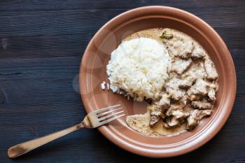 Russian cuisine dish - prepared portion of Beef Stroganoff (Beef Stroganov, Befstroganov) pieces of stewed meat in sour cream with boiled rice on brown plate on dark wooden board