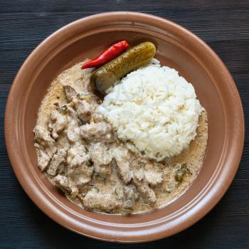 Russian cuisine dish - brown plate with Beef Stroganoff (Beef Stroganov, Befstroganov) pieces of stewed meat in sour cream and boiled rice dark wooden board