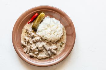 Russian cuisine dish - portion of Beef Stroganoff (Beef Stroganov, Befstroganov) pieces of stewed meat in sour cream with boiled rice on brown plate on white board with copyspace