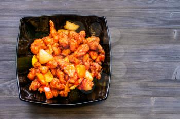 Chinese cuisine dish - top view of Fried chicken pieces with pineapple and bell pepper and onion (Sweet and Sour Chicken) in black bowl on dark wooden board with copyspace