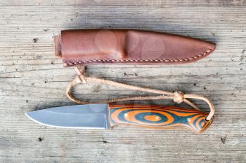 hand forged blackened steel knife with homemade epoxy and orange fabric handle with to hand-sewn leather sheath on old wooden board