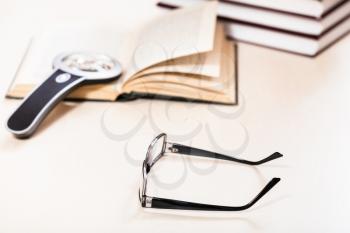 reading book with low vision - eyeglasses and magnifier on open book near stack of books on pale table (focus in the foreground)