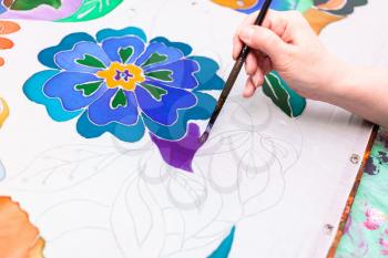 painter paints floral pattern on white silk canvas on wooden frame in cold contour batik technique