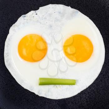 fried eggs and piece of celery on black plate close up. Fried eggs like face with straigh closed mouth