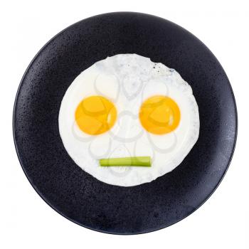 top view of fried eggs and piece of celery on black plate isolated on white background. Fried eggs like face with straigh closed mouth