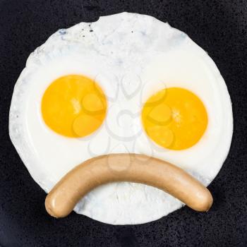 top view of fried eggs and curved boiled sausage on black plate close up. Fried eggs like frowning face