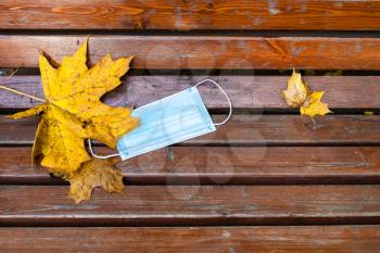 top view of thrown medical face fask and fallen maple leaves on wooden bench closeup on autumn day