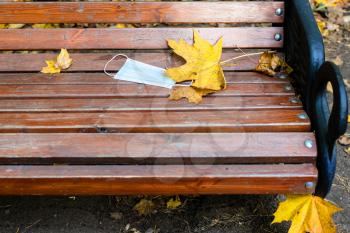 dropped medical face fask and yellow maple leaves on wooden bench in city park on autumn day