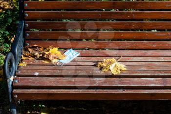 abandoned medical face fask and fallen maple leaves on wooden bench in city park on autumn day