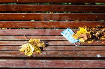 abandoned medical face fask and fallen maple leaves on surface of wooden bench closeup on autumn day