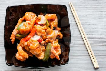 korean cuisine - top view of stir-fried Shrimps with cashew nuts and vegetables in sweet and sour sauce (Shrimps Combo) in black bowl on wooden table with chopsticks
