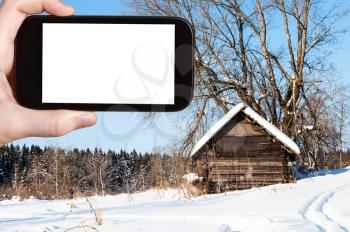 travel concept - tourist photographs of old abandoned wooden hut near forest in sunny winter day in little village in Russia on smartphone with cut out screen with blank place for advertising