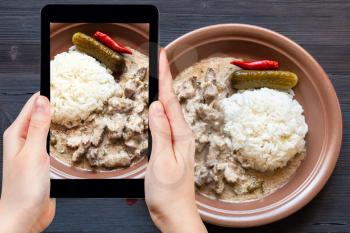 travel concept - visitor photographs of Russian cuisine dish of Beef Stroganoff (Beef Stroganov, Befstroganov) pieces of stewed meat in sour cream with boiled rice on smartphone