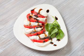 italian cuisine insalata caprese (caprese salad) - sliced mozzarella cheese and tomato with basil twig seasoned by olive oil and balsamic vinegar on white plate on wooden table