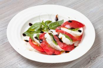 italian cuisine insalata caprese (caprese salad) - sliced mozzarella cheese and tomato with basil leaves seasoned by olive oil and balsamic vinegar on white plate on wooden table