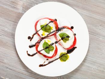 italian cuisine insalata caprese (caprese salad) - top view of stacks from sliced mozzarella cheese, tomato, basil leaf seasoned by olive oil, balsamic vinegar and pesto sauce on plate on wooden table