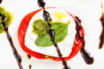 italian cuisine insalata caprese (caprese salad) - top view of stack from mozzarella cheese and tomato slices with basil leaf seasoned by olive oil, balsamic vinegar and pesto sauce on white plate