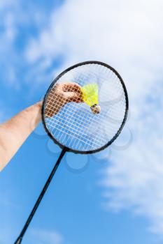 female hand strikes yellow shuttlecock by badminton racquet with background from blue sky with white clouds in sunny day (focus on the shuttlecock)