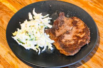 piece of roasted meat and cabbage and cucumber salad on black plate on wooden table at home kitchen