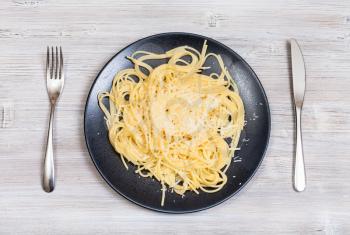 italian cuisine - top view of spaghetti al burro e parmigiano (pasta with butter and cheese) on black plate and fork with knife on gray wooden background