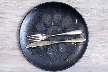 top view of knife and fork lie parallel on dirty black plate after lunch on gray wooden table