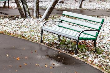 the first snow on wooden bench in city park on autumn day