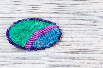 needlecraft background - top view of handcrafted oval brooch embroidered by various silk embroidery threads and violet beads on gray wooden board with copyspace