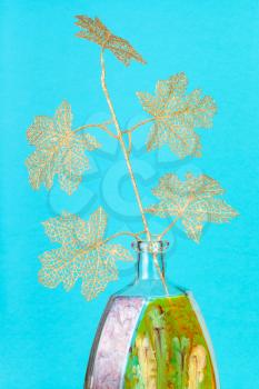 artificial golden plastic twig of tree in handpainted glass brandy bottle on aquamarine pastel color background