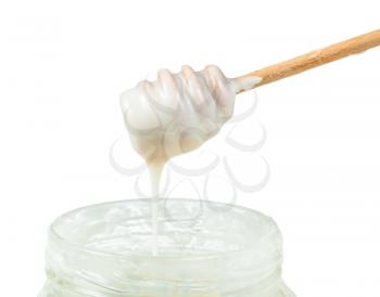 side view of natural organic white honey flows from wooden stick into glass jar closeup isolated on white background