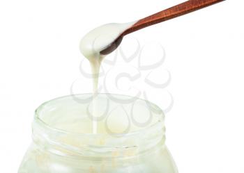 side view of natural organic white honey pouring from little wooden spoon into glass jar with honey closeup isolated on white background