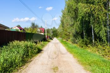 country road along green forest and fence of village in Russia on sunny summer day