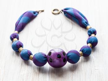 handcrafted necklace of round beads wrapped in blue silk cloth and purple plastic ball with holes on wooden table close up