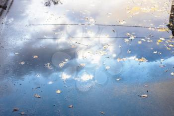 rain puddle on surface of asphalt footpath with fallen leaves and reflection of blue sky with clouds in city on autumn day