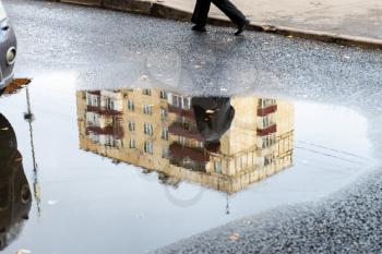 cityscape with rain puddle with reflection of municipal urban house and walking man on asphalt road on autumn day