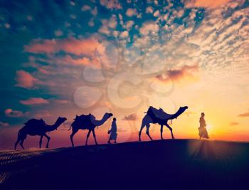 Vintage retro effect filtered hipster style image of Rajasthan travel background - two indian cameleers camel drivers with camels silhouettes in dunes of Thar desert on sunset. Jaisalmer, Rajasthan, I