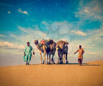 Vintage retro hipster style travel image of Rajasthan travel background - two Indian cameleers (camel drivers) with camels in dunes of Thar desert. Jaisalmer, Rajasthan, India with grunge texture over