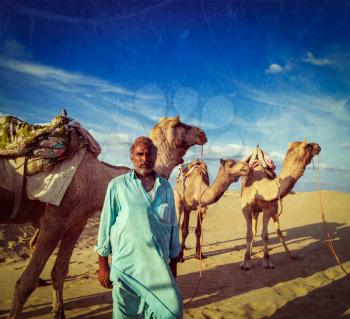 Vintage retro hipster style travel image of Rajasthan travel background - Indian man cameleer (camel driver) portrait with camels in dunes of Thar desert with grunge texture overlaid. Jaisalmer, Rajas