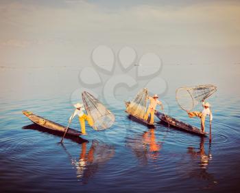Myanmar travel attraction - Traditional Burmese fishermen balancing with their fishing net on boats at Inle lake in Myanmar famous for their distinctive one legged rowing style. Vintage filtered retro