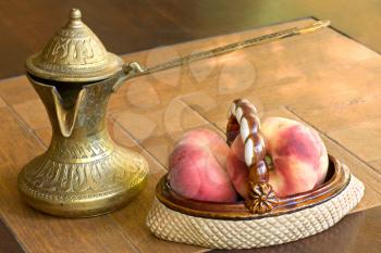Old brass arabian style coffee pot and ceramic vase with peaches on a table.